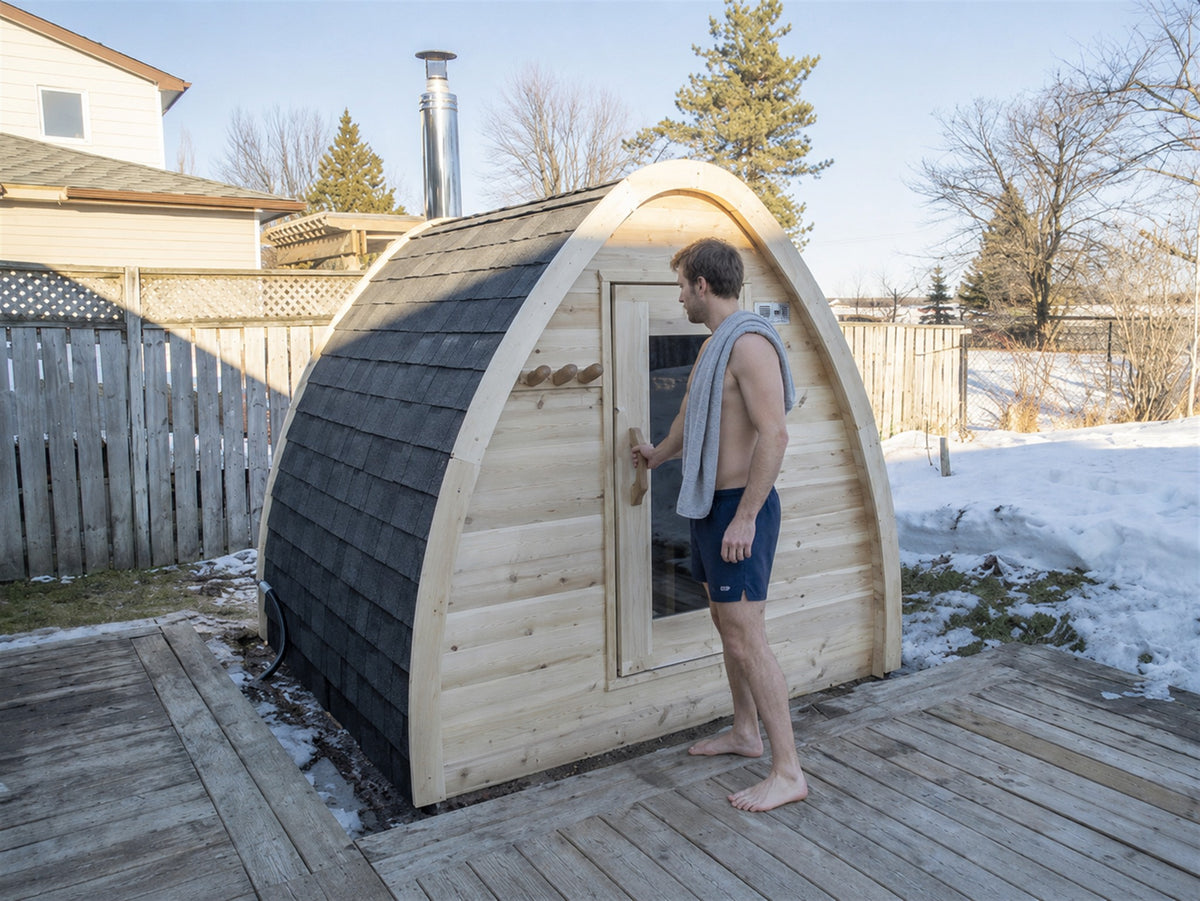 A man with a towel over his shoulder reaches for the door of the MiniPOD Sauna on a snowy backyard deck
